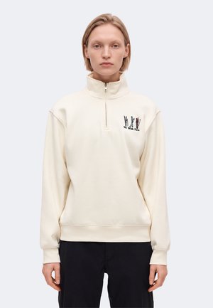 Modèle portant un sweat-shirt crème à fermeture éclair courte, avec un petit logo sur la poitrine et un pantalon noir, devant un fond uni clair.