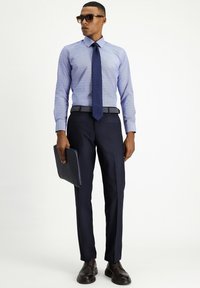 Lichtblauw blouse met patroon, marineblauwe stropdas, marineblauwe pantalon, gestructureerde riem, zwarte gestructureerde portefeuille, bruine leren schoenen en zonnebril.