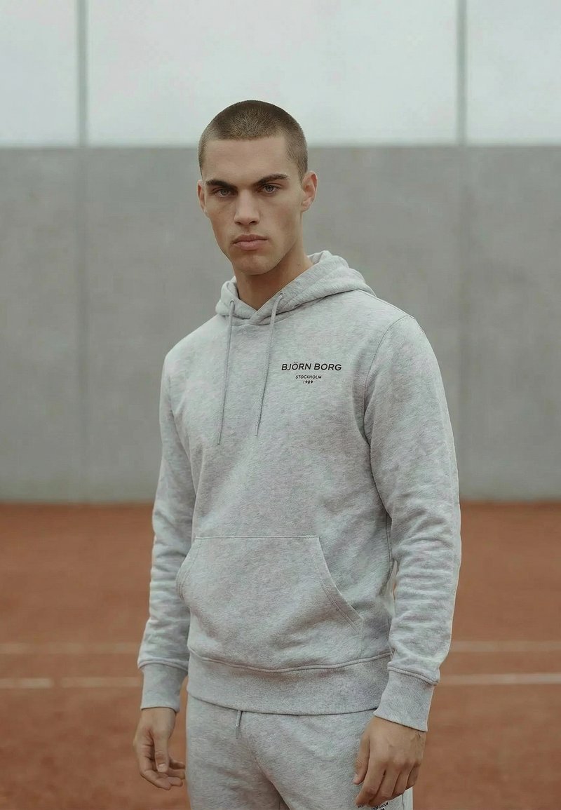 Joven con pelo corto que lleva una sudadera con capucha y pantalones Björn Borg de color gris claro, de pie en una pista de tierra batida roja con una pared gris como fondo.