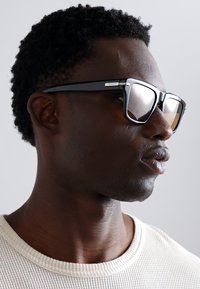 Lunettes de soleil avec une monture en plastique noir, des verres teintés de gris et des accents argentés sur les branches. Portées par une personne avec des cheveux courts et texturés.