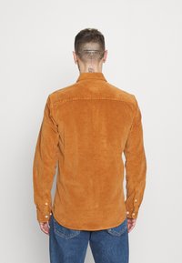 Mann mit kurzer Frisur und Ohrpiercings, der ein rotbraunes Cordhemd und blaue Jeans trägt und einer schlichten weißen Wand zugewandt ist.