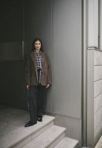 Bruine nubuck oversized blazer, zwart-wit geblokt shirt, zwarte wijde broek met witte zijkanten, en glanzende zwarte schoenen.