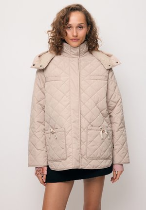 Kobieta o kręconych włosach, ubrana w beżową pikowaną kurtkę z wysokim kołnierzem, przednimi kieszeniami i różowymi dzianinowymi rękawami widocznymi pod spodem.