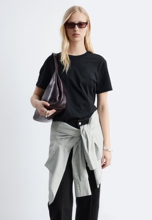 Blonde vrouw met een zwart T-shirt, zonnebril, een licht shirt om haar taille gestrikt en een donkere leren schoudertas vastgehouden.