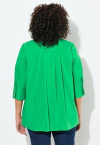 Camisa verde de manga larga con espalda plisada, que presenta mangas enrollables y una textura suave. Diseñada para ofrecer comodidad y facilidad de movimiento.
