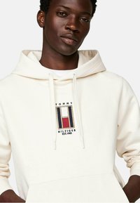 Sudadera color crema con cordones, bolsillo frontal y logo bordado de Tommy Hilfiger que presenta elementos en azul, rojo y blanco.