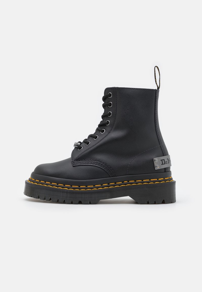 Botte de combat en cuir noir avec semelle épaisse, couture jaune, laçage à l'avant et languette de tirage de marque à l'arrière.