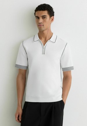 Férfi, aki fehér, rövid ujjú polo ingben áll, amelyen fekete szegély van a galléron, a cipzáron és az ujjvégeken, fekete nadrággal párosítva.