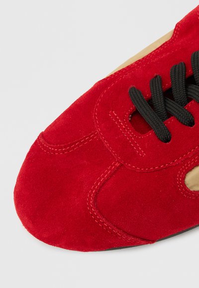 Μπροστινό μέρος κόκκινου παπουτσιού από σουέντ με μαύρα κορδόνια και ορατή ραφή σε ουδέτερο φόντο.