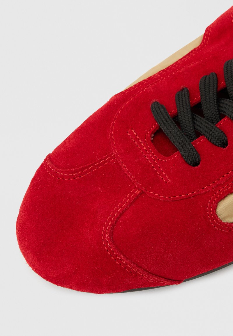 Parte frontal de zapato de ante rojo con cordones negros y costuras visibles sobre un fondo neutro.