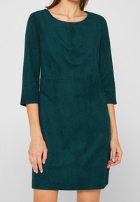 Robe bleu sarcelle en velours jusqu'aux genoux avec des manches trois-quarts et un col rond, portée par une femme avec les bras détendus le long du corps.