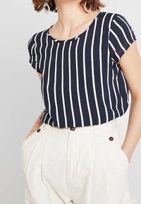 Top a righe blu navy e bianche con maniche corte e scollatura rotonda, realizzato in un tessuto morbido; abbinato a pantaloni bianchi a vita alta.