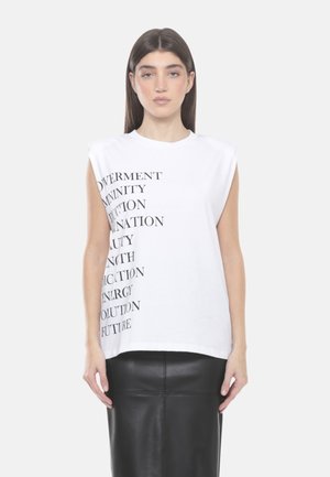 Donna con lunghi capelli scuri indossa un top bianco senza maniche con testo nero e una gonna in pelle nera, in piedi contro uno sfondo chiaro e semplice.