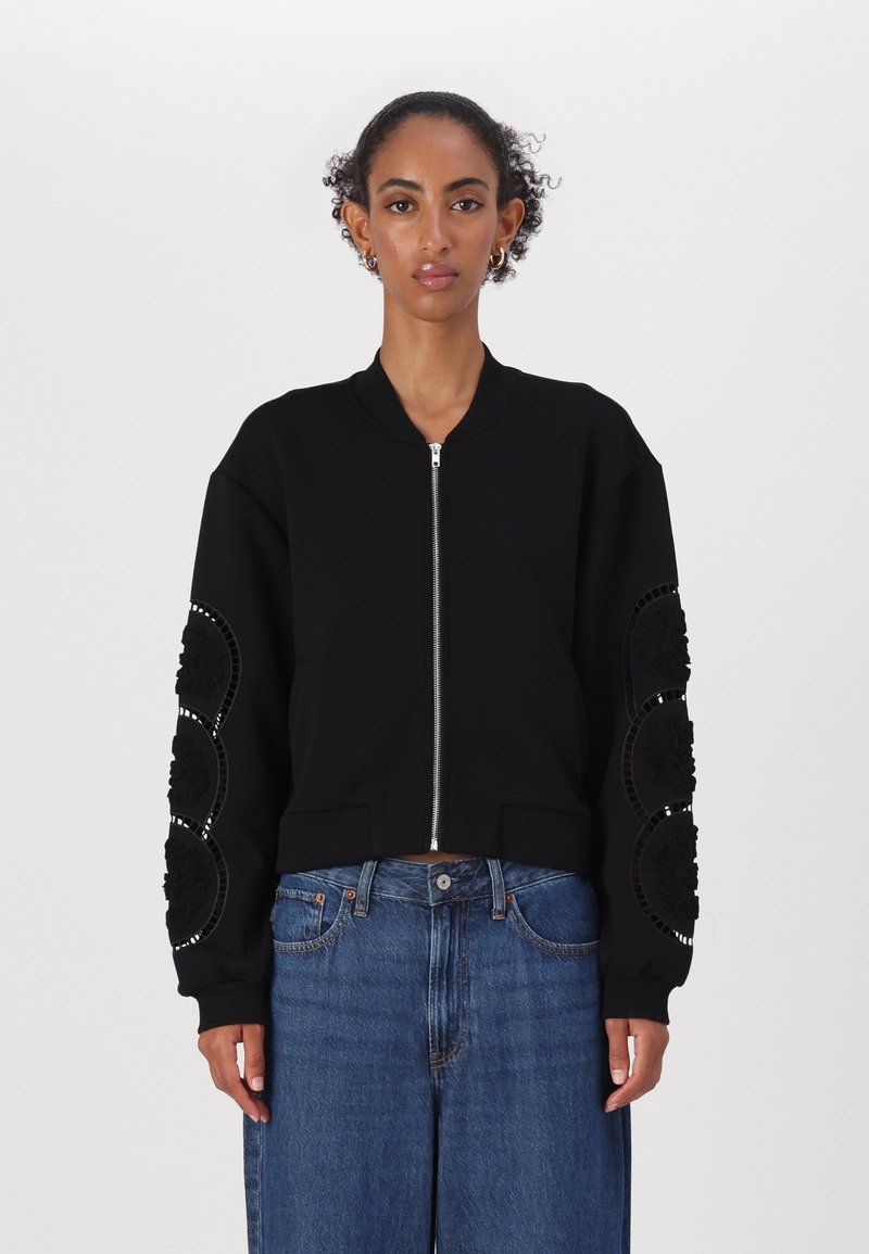 Čierna bomber bunda so zapínaním na zips, ribbed lemom a textúrovanými kvetinovými detailmi na rukávoch. Kombinovaná s modrými džínsami.