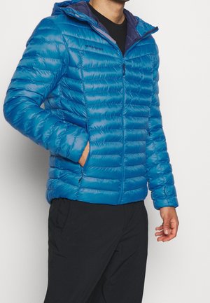 Man wearing a blue quilted Mammut jacket with hood, black pants, left hand in pocket, standing against a plain background.