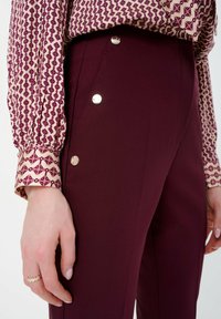 Pantalon bordeaux au toucher lisse, agrémenté de trois boutons argentés sur le côté, associé à un chemisier à motifs crème et marron.