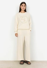 Beige sweatshirt met een cirkelvormig "Life" logo, gecombineerd met een wijde beige broek. Zachte stoftextuur, gedragen met donkere slippers.