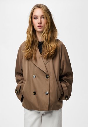 Jeune femme aux longs cheveux blonds portant une veste marron ample à double boutonnage avec des boutons et un pantalon blanc, les mains dans les poches.
