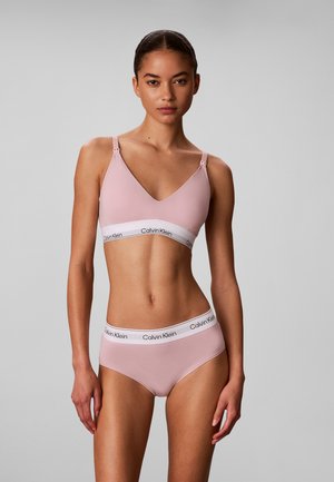 Femme portant un bralette rose clair Calvin Klein et une culotte assortie, debout devant un fond gris uni, regardant droit devant avec une expression neutre.