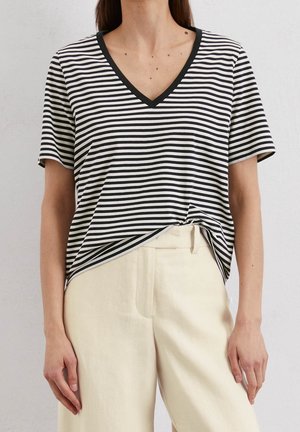 Kobieta ubrana w czarno-biały paskowany T-shirt z krótkim rękawem i dekoltem w serek, wpuszczony do beżowych spodni z wysokim stanem, stojąca na jednolitym tle.