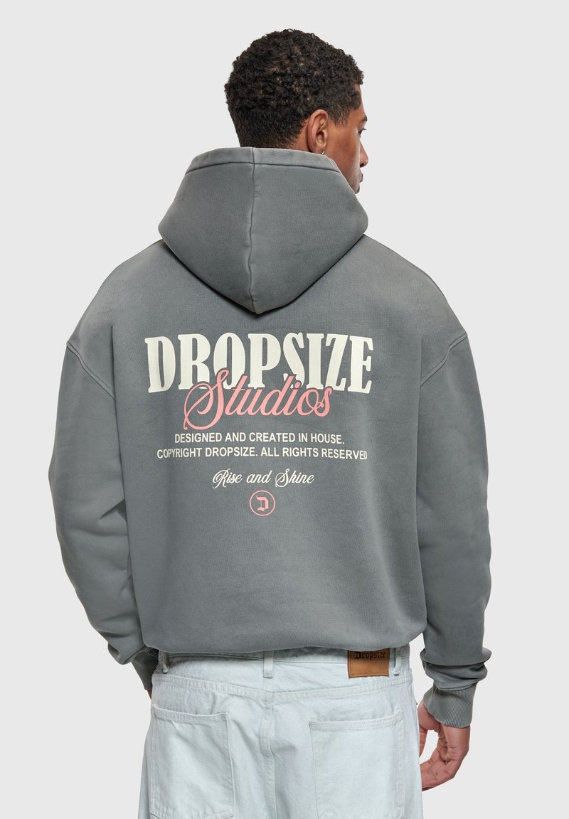 Homme de dos portant un sweat à capuche gris avec les inscriptions « Dropsize Studios » et « Rise and Shine » au dos, associé à un pantalon bleu clair.