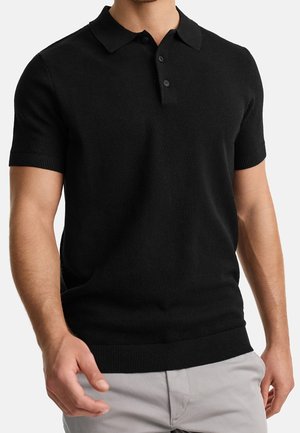 Homme portant un polo en maille noir à manches courtes et un pantalon gris clair, debout avec les bras détendus devant un fond uni.