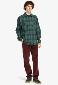 Grøn flannel skjorte med et ternet mønster, knaplukning fortil og to brystlommer, parret med bordeaux kordfløjl bukser og mørke sneakers.