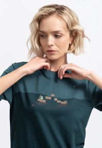 Donker teal katoen T-shirt met een landschapafbeelding van boten en heuvels in gedempte kleuren. Korte mouwen met een klassieke ronde hals.
