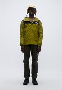 Πράσινο αντιανεμικό μπουφάν με μαύρες λεπτομέρειες, σε συνδυασμό με σκούρο πράσινο παντελόνι και ανοιχτόκίτρινο καπέλο τύπου κάπα. Μαύρα παπούτσια. Ομοιόμορφο φόντο.