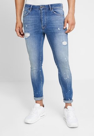 Slim-fit blauwe spijkerbroek met opgerolde boorden, versleten plekken en verfspatten, gedragen met witte sneakers door een staande persoon.
