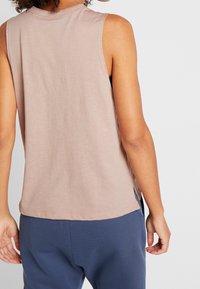 Top senza maniche di un delicato rosa chiaro, realizzato in un tessuto morbido, con scollatura rotonda e fessure laterali per il comfort. Indossato con pantaloni blu navy.