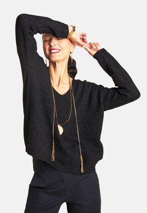 Schwarzer gestrickter Pullover mit V-Ausschnitt, weiten Ärmeln und lockerem Schnitt. Beinhaltet eine lange goldene Kette mit Quasten. Glatte Textur, leicht schimmernd.