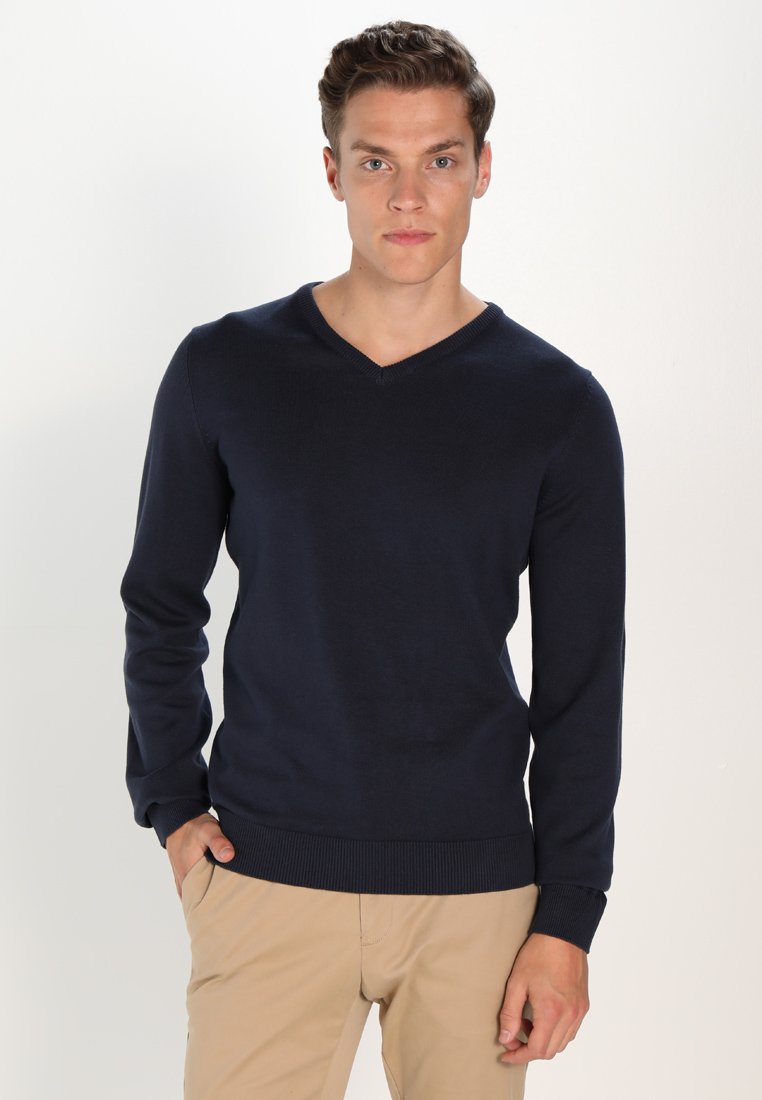 Navyblauer V-Ausschnitt-Pullover aus weichem Strickstoff, mit langen Ärmeln und geripptem Saum. Das Model trägt ihn mit hellbeigen Hosen.