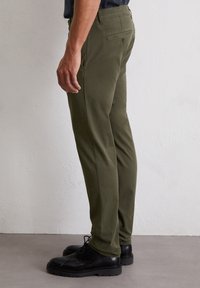 Olivgrüne Hosen mit geradem Schnitt, aus glattem Stoff gefertigt; mit einer Gesäßtasche und Gürtelschlaufen versehen; kombiniert mit schwarzen Schnürschuhen.