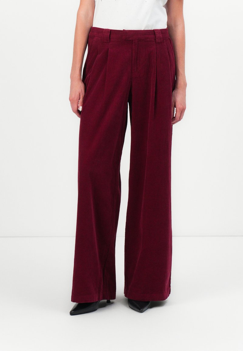 Pantalons larges bordeaux en velours côtelé ; avec des plis à l'avant, une taille haute et une texture lisse. Associés à des talons noirs.