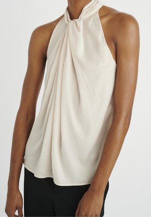 Sleeveless light beige halter top with twisted neckline and draped front worn with black pants on a person with medium brown skin.