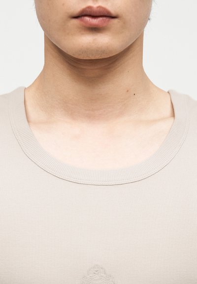 Κοντινό πλάνο του κάτω μέρους του προσώπου, του λαιμού και του ανώτερου στήθους ενός ατόμου που φοράει μπεζ ριγωτό τοπ με στρογγυλή λαιμόκοψη.