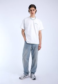 Vit bomull T-shirt med en liten grön grafisk design, i kombination med ljusblå jeans och silvriga löparskor. Modellen bär ett färgglatt pärlhalsband.