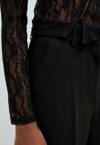 Haut en dentelle noire à manches longues avec des motifs floraux, associé à un pantalon noir ajusté. Le haut présente une texture transparente et des détails de boutons.