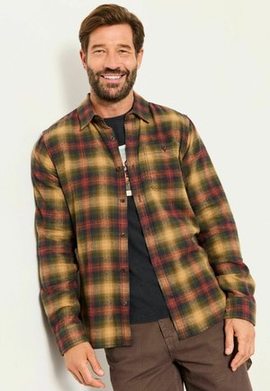 Lächelnder Mann mit Bart, der ein gelb-grün-rotes kariertes Hemd über einem schwarzen T-Shirt trägt und vor einem schlichten hellen Hintergrund steht.
