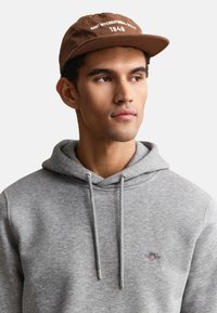 Jeune homme portant une casquette marron avec un texte blanc et un sweat à capuche gris clair avec un petit logo rouge et bleu sur la poitrine, regardant sur le côté.