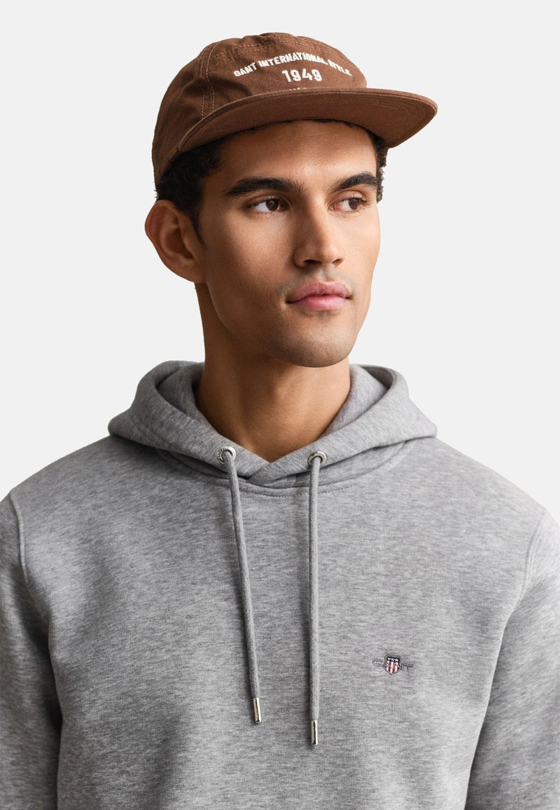 Joven con gorra marrón con texto blanco y sudadera gris claro con un pequeño logo rojo y azul en el pecho, mirando hacia un lado.