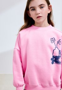 Pink sweatshirt med et grafisk tryk af en vase med blomster. Den har rund halsudskæring, sænkede skuldre og ærmer med manchetter.