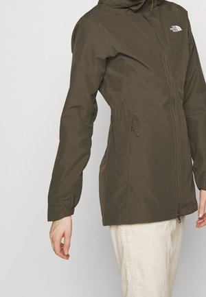 Person wearing olive green North Face zip-up jacket with elastic cuffs and light-colored pants against neutral background.