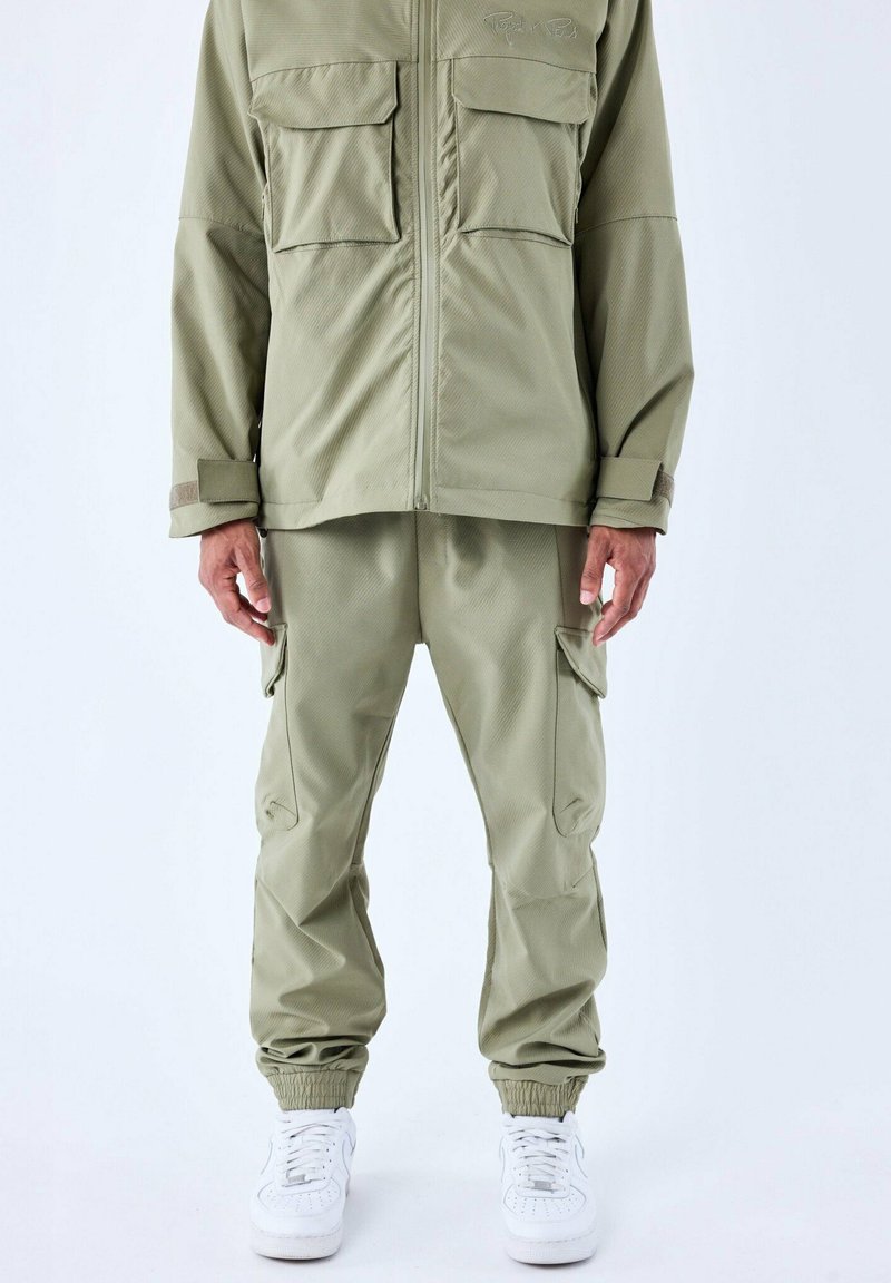 Olijfgroene jas met twee borstzakken en bijpassende cargobroek. Beide hebben een zachte textuur en elastische manchetten. Gemaakt met witte sneakers.
