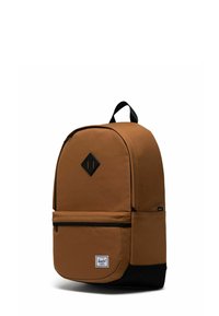 Sac à dos en tissu marron avec une forme arrondie, des accents noirs, une poche avant zippée et un écusson logo en forme de diamant à l'avant. Bretelles ajustables.