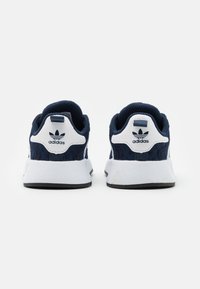 Pari tummansinisistä ja valkoisista Adidas-lenkkareista takaviistosta, joissa on valkoiset välikerrokset ja Adidas-logo kantapaikoissa.