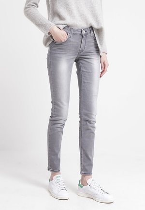 Osoba nosząca jasnoszare jeansy typu slim-fit, miękki szary sweter i białe trampki z zielonymi akcentami, stojąca z jedną ręką w kieszeni.