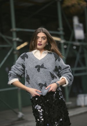 Grijze, oversized trui met zwarte pailletten vlinderpatronen, gedragen over een witte blouse met kraag; in combinatie met een glanzende zwarte pailletten rok.