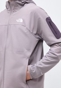 Άτομο που φοράει ανοιχτό μωβ ζακέτα North Face με φερμουάρ και τσέπη με φερμουάρ στο πάνω μέρος του μπράτσου, με το χέρι στην πλαϊνή τσέπη.